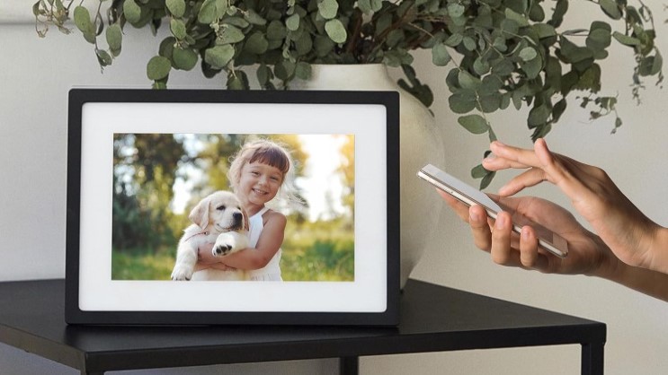 En hand använder mobilen för att justera bilderna på en fotoram som står på ett litet bord.